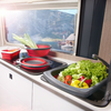Évier avec une bassine grise pliable contenant de la laitue, des poivrons jaune et rouge et des tomates cerises ; casseroles rouges pliables sur la plaque ; fenêtre donnant sur des montagnes et de l'eau