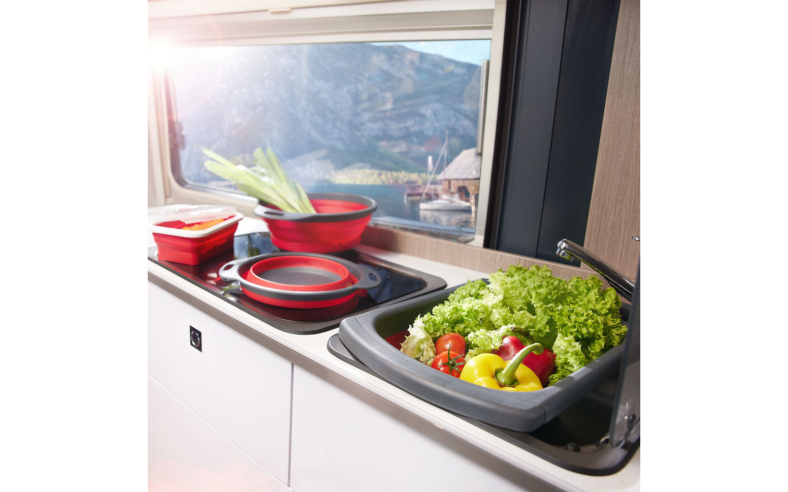Évier avec une bassine grise pliable contenant de la laitue, des poivrons jaune et rouge et des tomates cerises ; casseroles rouges pliables sur la plaque ; fenêtre donnant sur des montagnes et de l'eau
