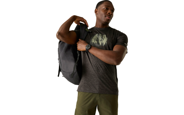 Person trägt schwarzen Rucksack über eine Schulter, dunkles T‑Shirt mit Aufdruck, olivgrüne Shorts und Armbanduhr, schaut nach rechts vor hellem Hintergrund