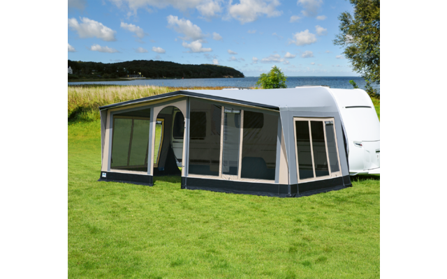 Weißer Wohnwagen mit angebautem Zelt auf grünem Gras vor See und blauem Himmel
