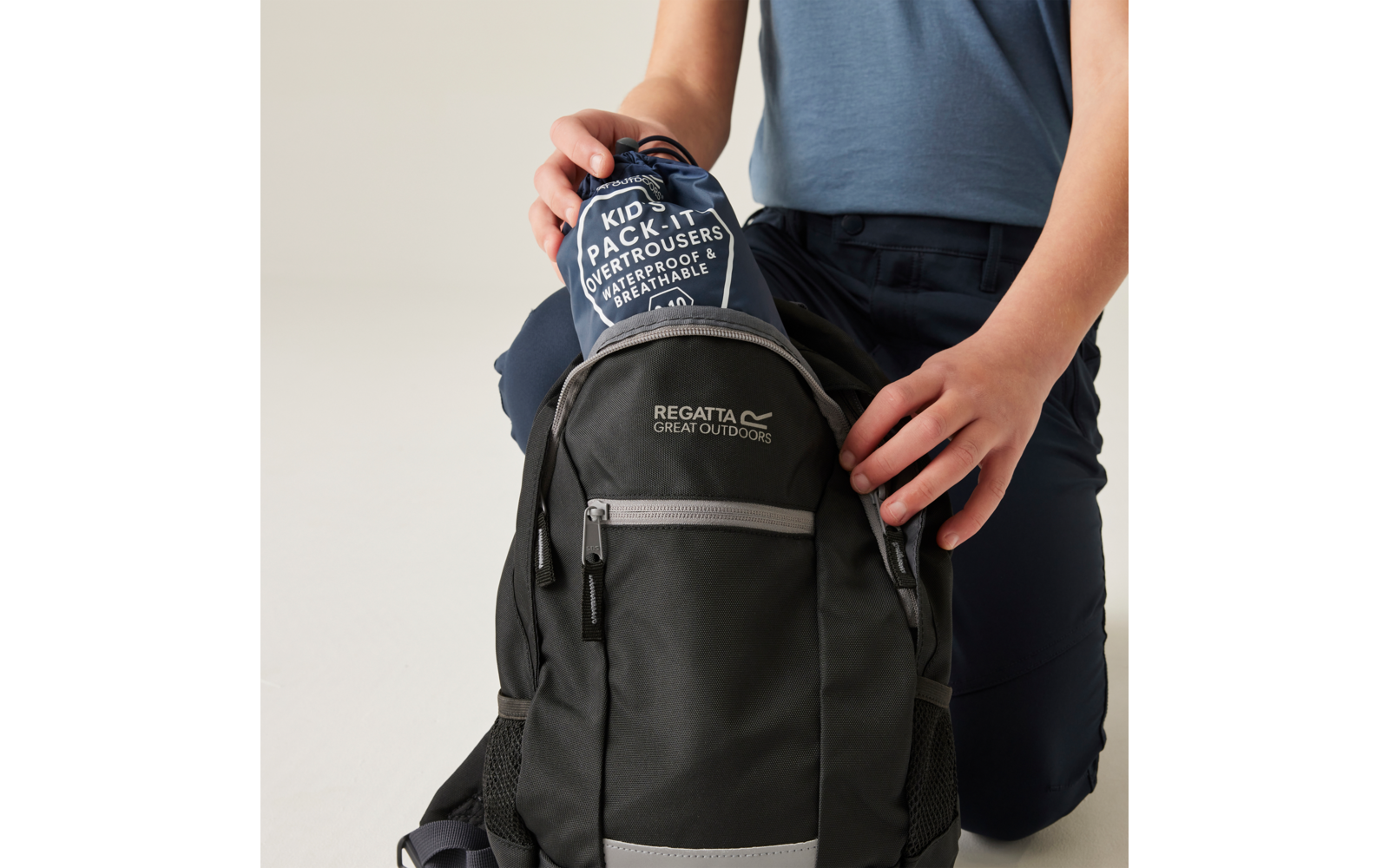 Handen en bovenlichaam van een persoon in een blauw bovenstuk die een verpakte broek met opschrift 'KID PACK IT OVERTROUSERS' in een zwarte rugzak met het logo 'REGATTA GREAT OUTDOORS' stopt