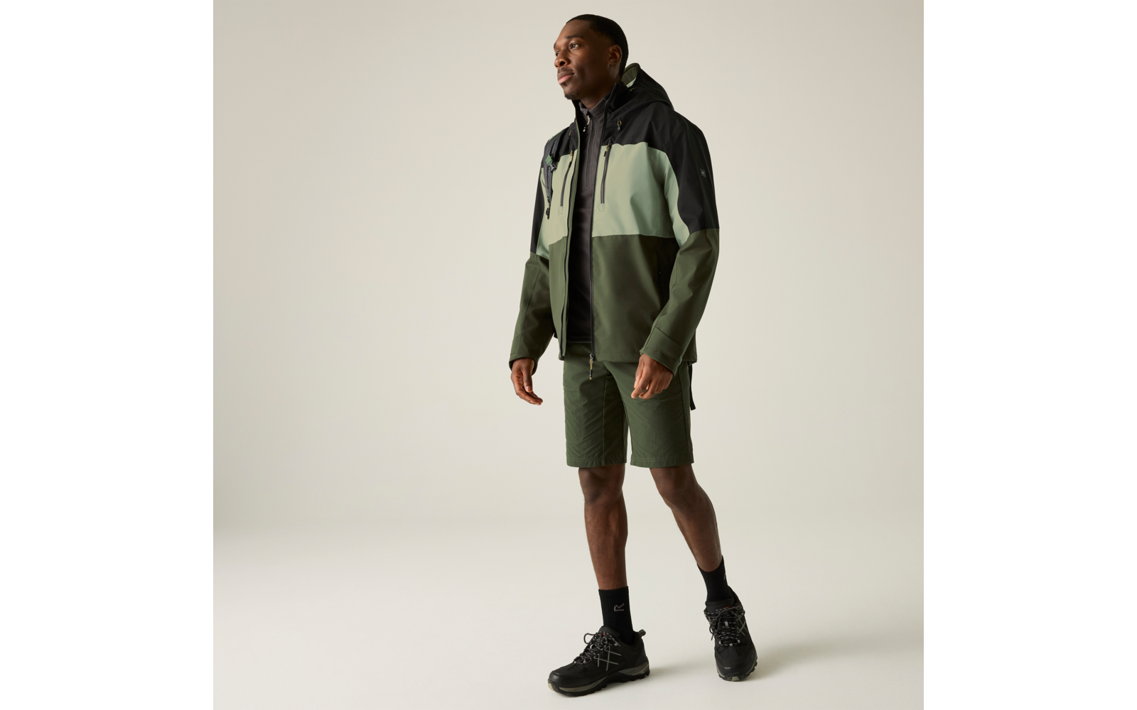 Person mit Kapuzenjacke (hellgrün, dunkelgrün, schwarz), olivgrüne Shorts, schwarze Socken und schwarze Schnürschuhe, steht vor hellem einfarbigem Hintergrund und blickt zur Seite