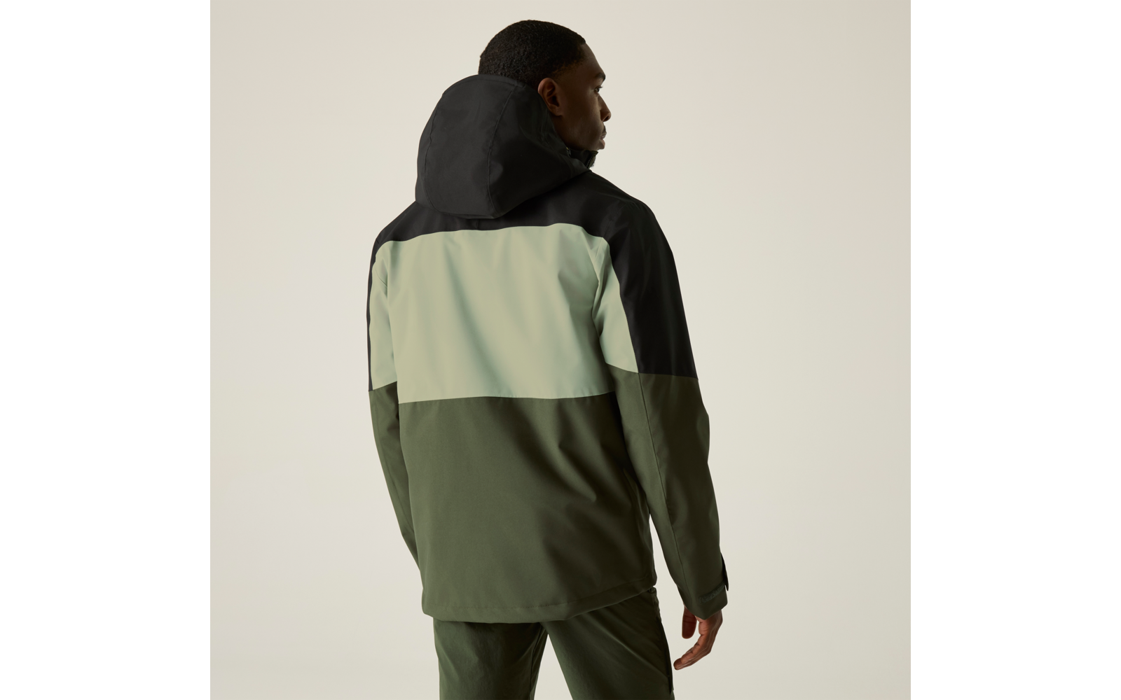 Person von hinten, trägt eine Kapuzenjacke mit schwarzer Kapuze, hellgrünem Mittelteil und olivgrünem Unterteil, vor beigem Hintergrund