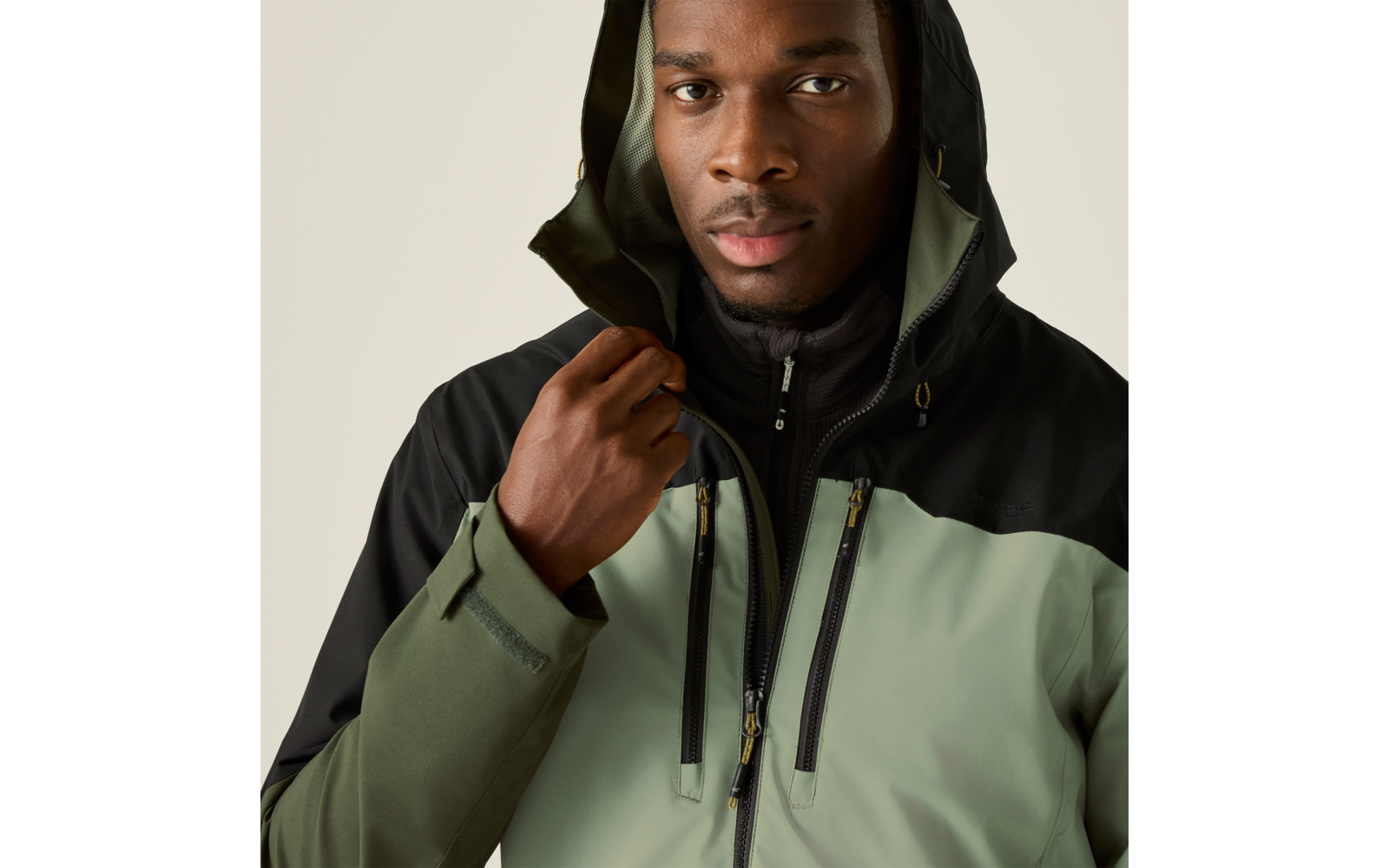 Person trägt grün-schwarze Kapuzenjacke, hält den Kragen, sichtbare Reißverschlüsse und Brusttaschen, heller einfarbiger Hintergrund