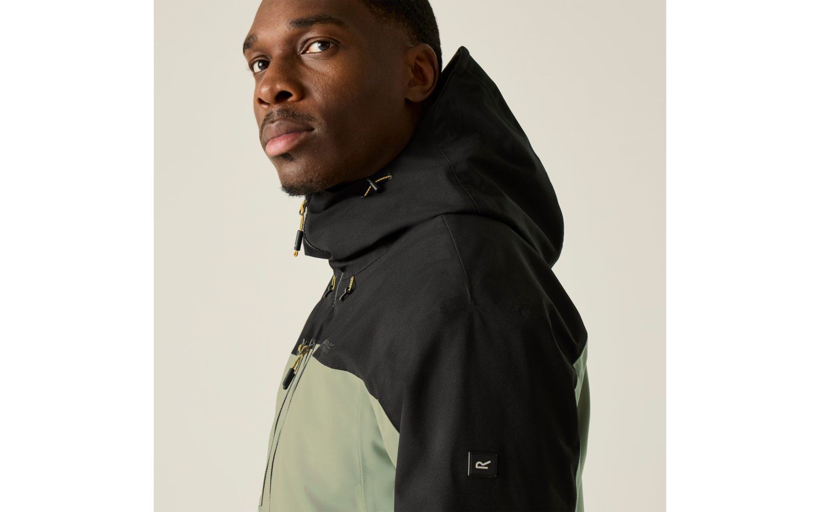 Person in Dreiviertelansicht trägt eine zweifarbige Kapuzenjacke (schwarze Kapuze, hellgrüner Oberkörper), Blick nach links, heller einfarbiger Hintergrund