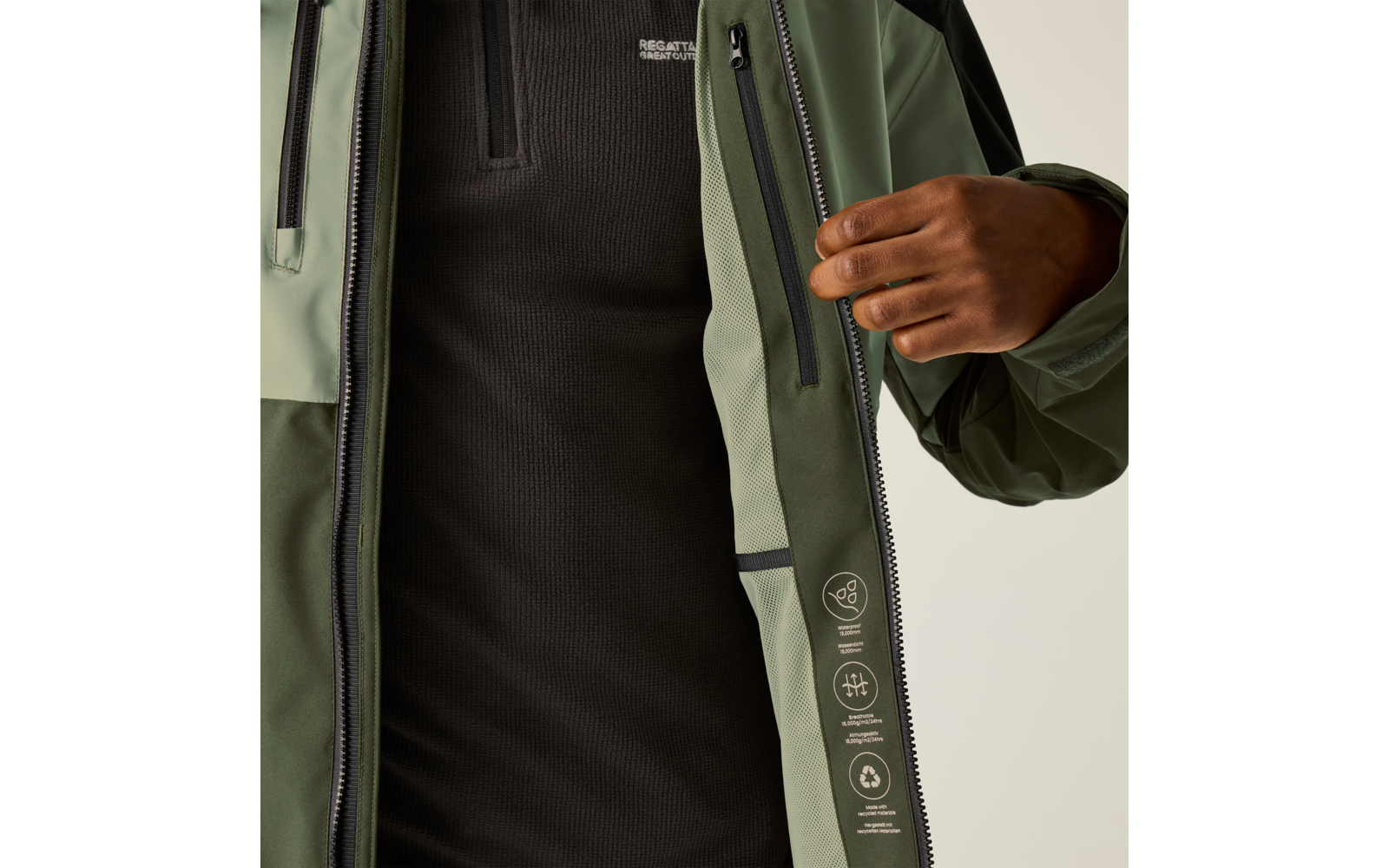 Nahaufnahme einer geöffneten olivgrünen Jacke mit Reißverschluss; rechte Hand hält die Innenkante; darunter ein schwarzes Oberteil mit kleinem Brustlogo
