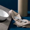 White faceted bowl on a dark base on a stone-colored slab, beige napkin with two white spoons, tall beige cylindrical vase in the background, blue surface and dark blue background