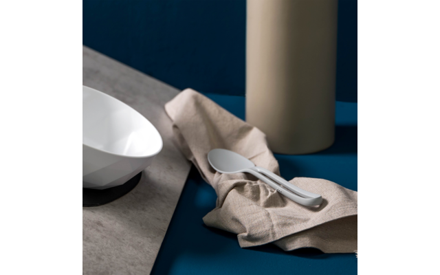 White faceted bowl on a dark base on a stone-colored slab, beige napkin with two white spoons, tall beige cylindrical vase in the background, blue surface and dark blue background