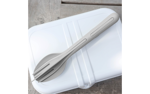 White rectangular plastic box with closed lid on a wooden surface; a light gray utensil with fork tines and a spoon bowl lies diagonally on the lid