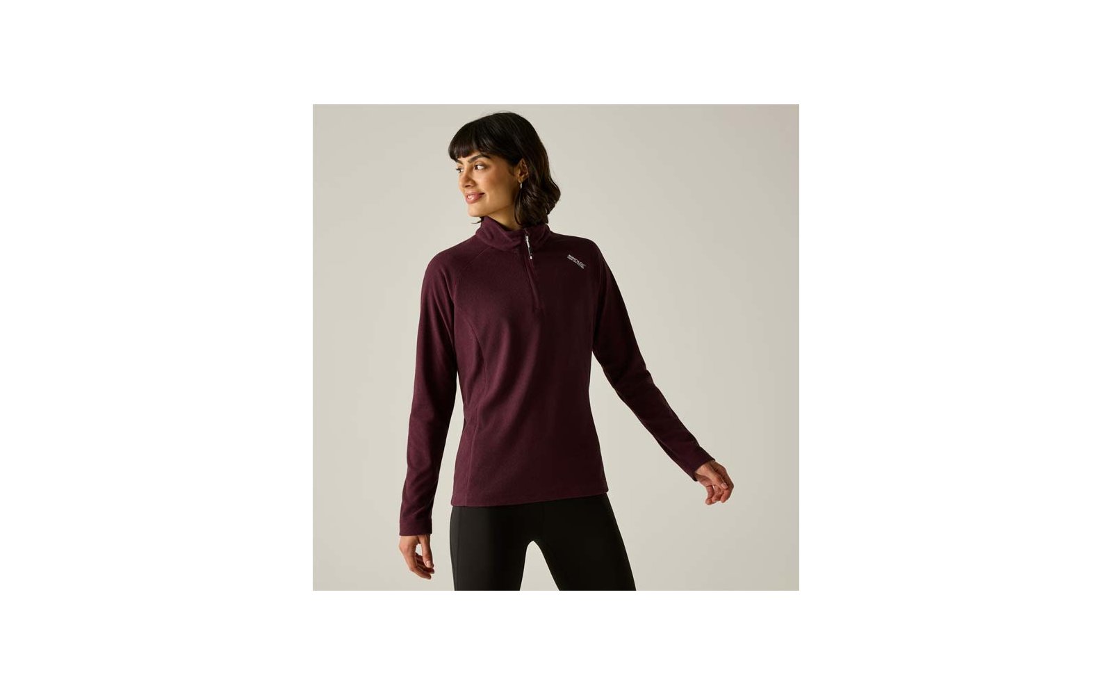 Personne devant un fond gris clair portant un pull bordeaux à manches longues avec une fermeture éclair courte et un petit logo sur la poitrine gauche, legging noir, regard vers la gauche