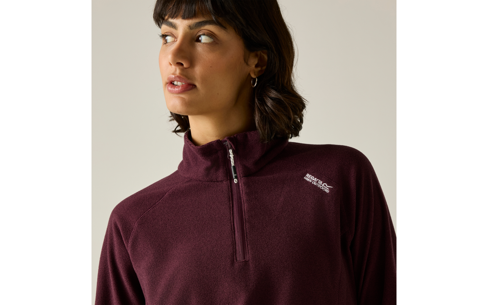 Personne aux cheveux foncés mi-longs et boucle d'oreille créole, regard vers la gauche ; porte un pull polaire bordeaux avec une petite fermeture éclair et un petit logo blanc, fond clair
