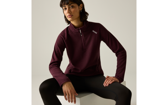 Personne assise sur un cube blanc, porte un pull bordeaux à demi-fermeture éclair, legging noir, cheveux courts foncés, clous d'oreille, arrière-plan clair neutre