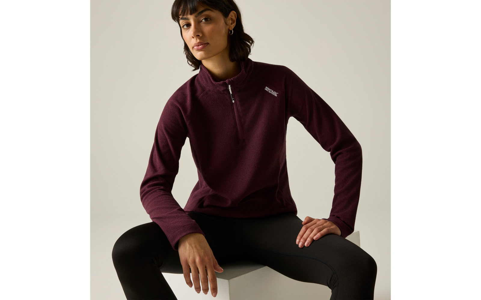Personne assise sur un cube blanc, porte un pull bordeaux à demi-fermeture éclair, legging noir, cheveux courts foncés, clous d'oreille, arrière-plan clair neutre