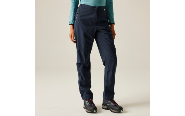 Person von der Taille abwärts in dunkelblauen Outdoor-Hosen, türkisfarbenem Langarmshirt und schwarzen Wanderschuhen vor hellem Hintergrund