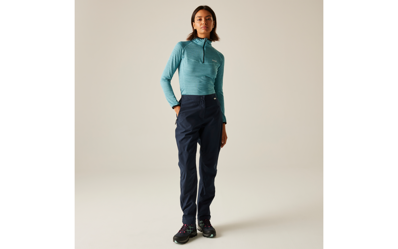 Frau steht vor hellem Hintergrund, trägt türkisfarbenes Langarmoberteil mit Reißverschluss, dunkelblaue Hose und schwarze Schuhe, eine Hand in der Hosentasche