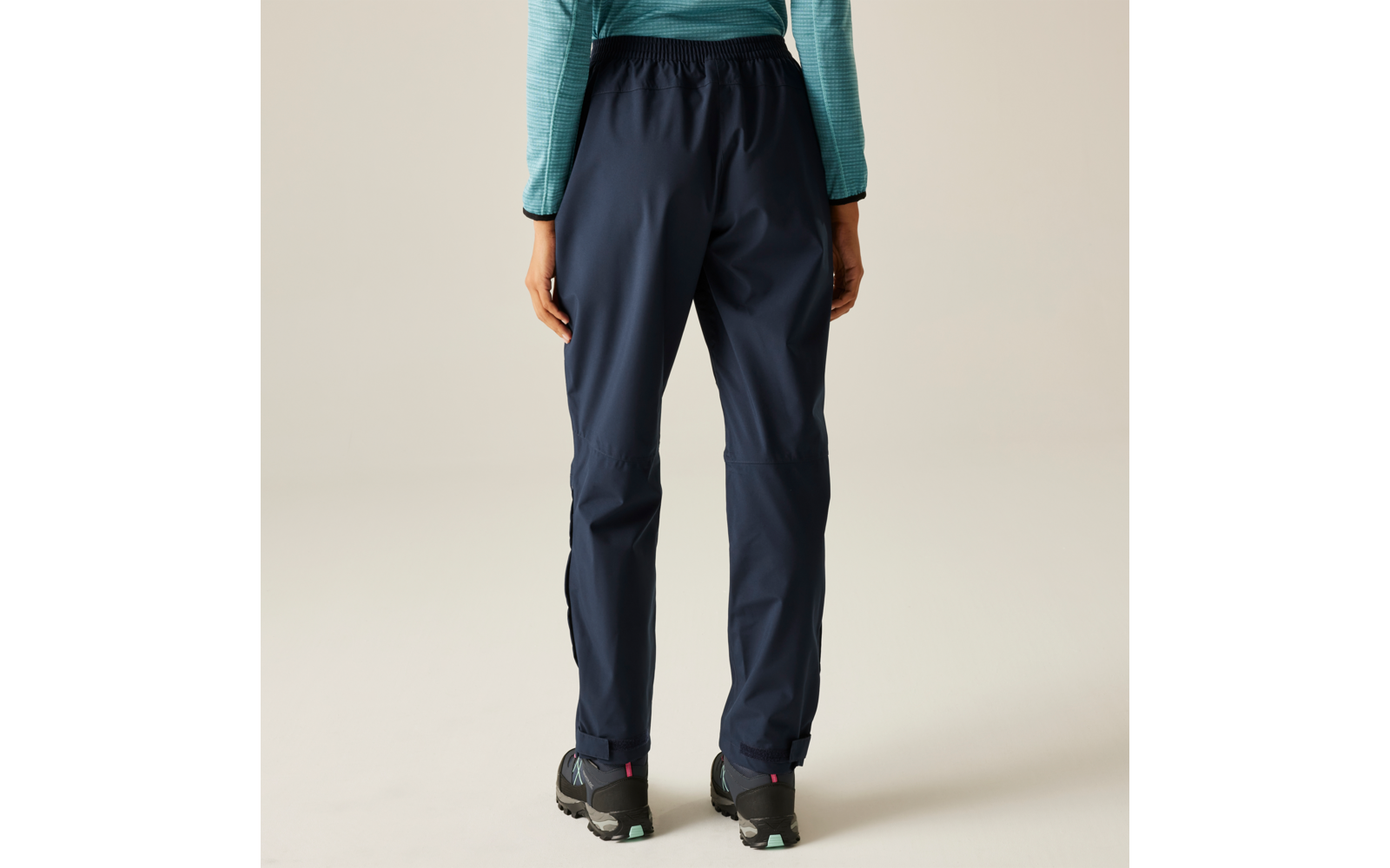 Rückenansicht einer Person mit dunkelblauen Hosen, türkisfarbenem Langarmoberteil und schwarzen Wanderschuhen vor hellem Hintergrund