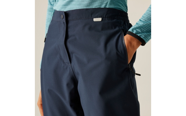 Nahaufnahme einer Person von Taille bis Hüfte, dunkelblaue Hose mit Hand in seitlicher Reißverschlusstasche und türkisfarbenes Langarmshirt