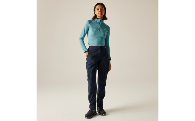 Frau steht vor hellem Hintergrund, trägt türkisfarbenes Langarmoberteil mit Reißverschluss, dunkelblaue Hose und schwarze Schuhe, eine Hand in der Hosentasche