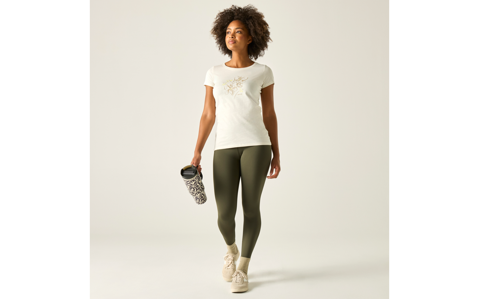 Personne aux cheveux bouclés portant un t-shirt blanc à manches courtes avec motif floral, leggings vert olive et baskets beige, tenant une bouteille à motif dans la main droite, marchant devant un fond clair uni