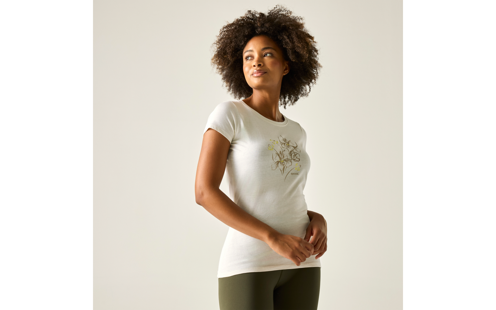 Personne aux cheveux bouclés portant un t‑shirt blanc à motif floral et des leggings olive, debout de profil léger devant un fond clair et regardant en haut à droite