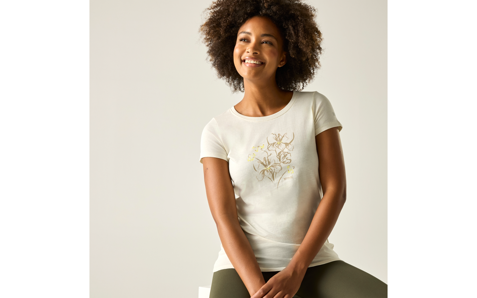 Personne assise sur un tabouret, sourit, porte un T-shirt clair à motif floral et un pantalon vert olive, cheveux bouclés, fond uni et clair