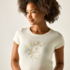 Femme aux cheveux bouclés, demi‑portrait ; elle porte un t‑shirt clair à motif floral, regarde sur le côté et sourit légèrement sur fond clair