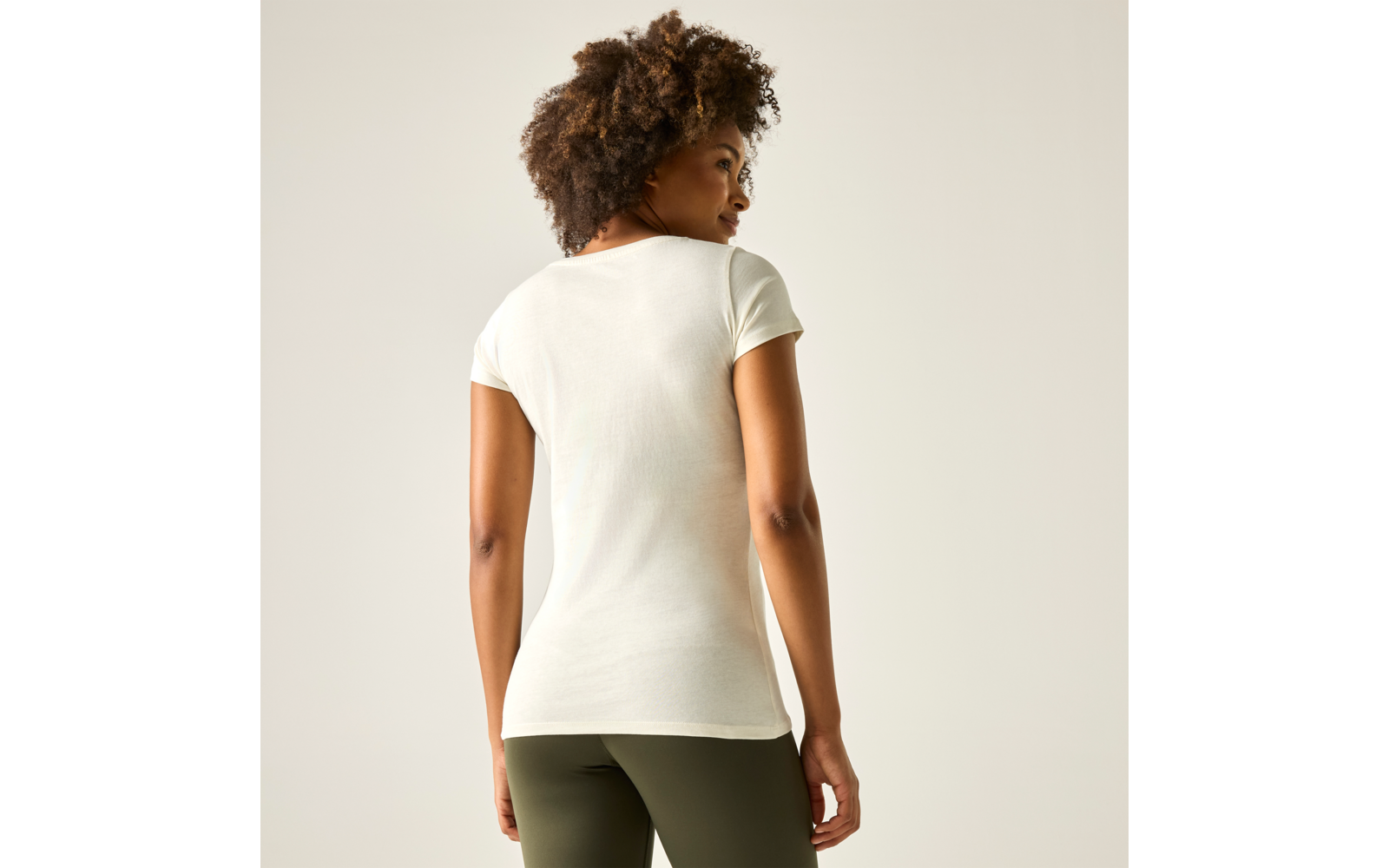 Personne de dos, debout devant un fond clair, porte un t‑shirt blanc à manches courtes et des leggings vert olive, regarde par-dessus son épaule droite, cheveux courts et bouclés