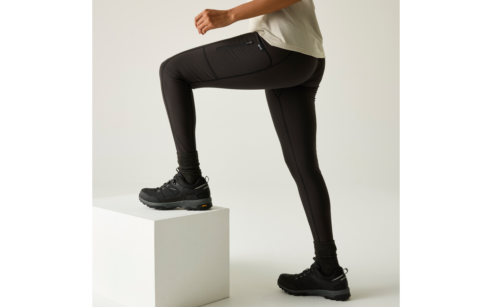 Unterkörper einer Person, ein Fuß auf einem weißen Block, schwarze Wanderschuhe, schwarze Leggings, Saum eines hellen Shirts sichtbar, heller einfarbiger Hintergrund