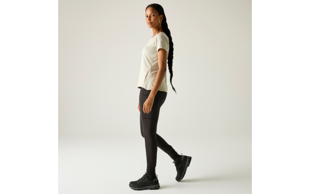 Seitlich stehende Person mit langem geflochtenem Zopf, hellem T‑Shirt, dunklen Leggings und schwarzen Sneakers vor hellem Hintergrund