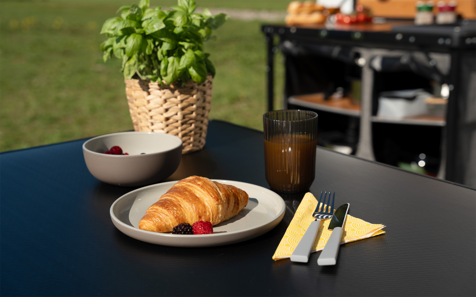 Tisch mit Croissant auf weißem Teller, zwei rote Beeren daneben, weiße Schale mit roten Beeren, hohes Glas mit orangefarbenem Getränk, Messer und Gabel auf gelbem Serviette, geflochtener Topf mit Basilikum, schwarze Tischfläche, verschwommener Küchen