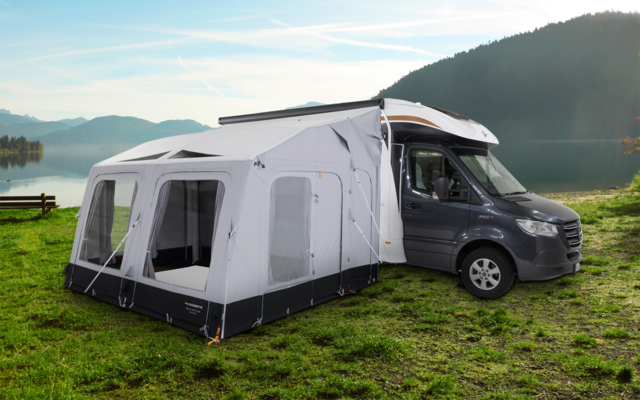 Een camper met een aangebouwde luifel staat op een grasveld voor een meer en bergen