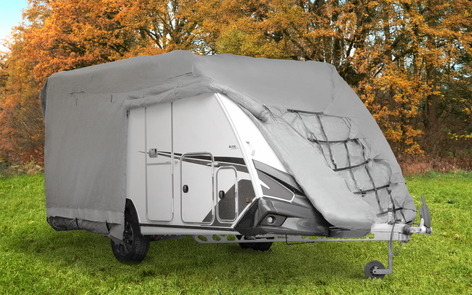 Weißer Wohnwagen, teilweise von einer grauen Abdeckplane bedeckt, auf grüner Wiese; im Hintergrund Bäume mit orangefarbenen und gelben Blättern; sichtbare Anhängerkupplung und vorderes Seitenfach