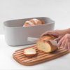 Ein Brotmesser schneidet ein Brot auf einem Holzbrett, neben einem grauen Behälter mit Croissants