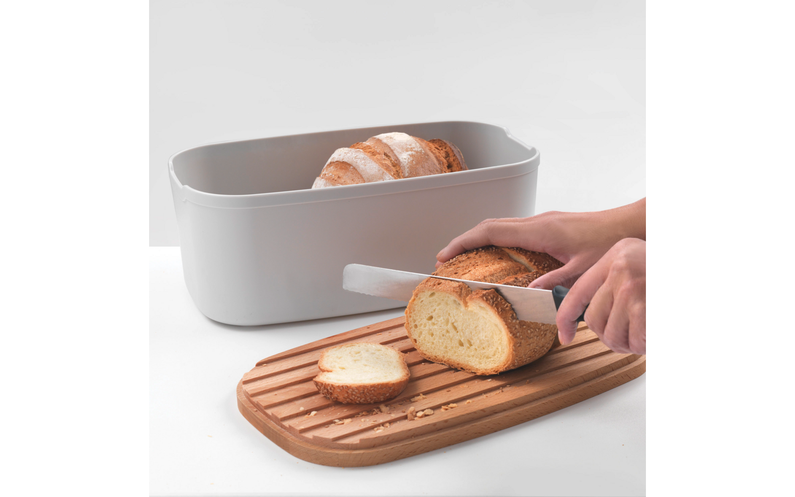 Ein Brotmesser schneidet ein Brot auf einem Holzbrett, neben einem grauen Behälter mit Croissants