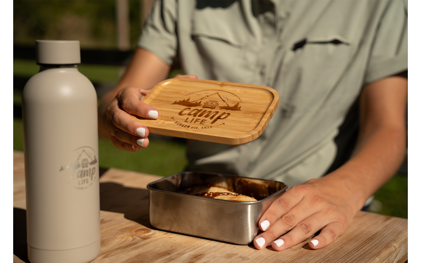 Hands with white nail polish holding a wooden lid engraved with a mountain logo and the text 'camp life' above a stainless‑steel lunchbox with a sandwich; left a gray water bottle with the same logo; wooden table; blurred green background