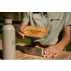 Mains avec vernis blanc tenant un couvercle en bois gravé d'un logo montagne et du texte 'camp life' au‑dessus d'une boîte à lunch en acier inoxydable contenant un sandwich ; à gauche une bouteille grise avec le même logo ; table en bois ; arrière‑plan vert flou