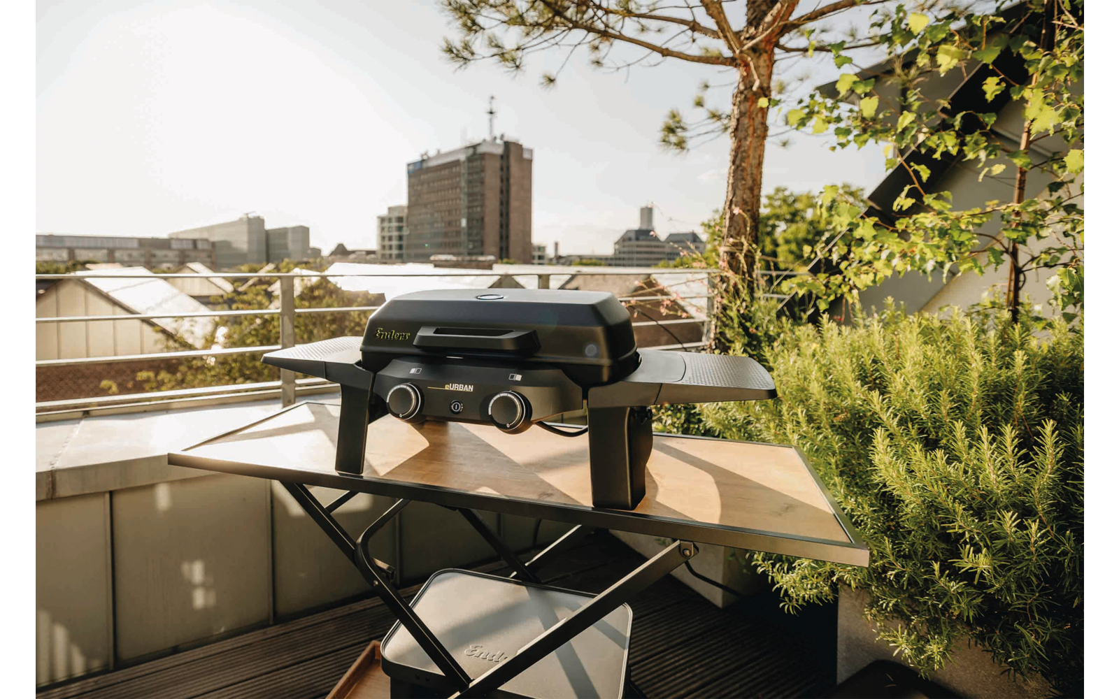 Kleiner Tischgrill auf einem Klapptisch auf einem Balkon, Pflanzen und Geländer im Vordergrund, Stadtgebäude und Himmel im Hintergrund