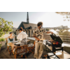 Dachterrasse mit drei Personen, eine Person steht am Grill, zwei sitzen an einem gedeckten Holztisch, Stadtdächer im Hintergrund