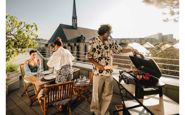Dachterrasse mit drei Personen, eine Person steht am Grill, zwei sitzen an einem gedeckten Holztisch, Stadtdächer im Hintergrund