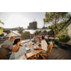Vier Personen sitzen an einem Holztisch auf einer Dachterrasse; Tisch mit Tellern, Gläsern und Flaschen; im Hintergrund Stadtgebäude, rechts ein Baum und Pflanzen, links Geländer