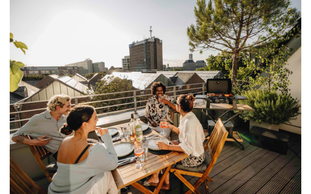 Vier Personen sitzen an einem Holztisch auf einer Dachterrasse; Tisch mit Tellern, Gläsern und Flaschen; im Hintergrund Stadtgebäude, rechts ein Baum und Pflanzen, links Geländer