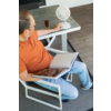 Personne assise sur une chaise extérieure avec haut orange et jean, tenant un livre ouvert sur les genoux ; sur la table à côté un petit ventilateur blanc et un verre contenant une boisson foncée