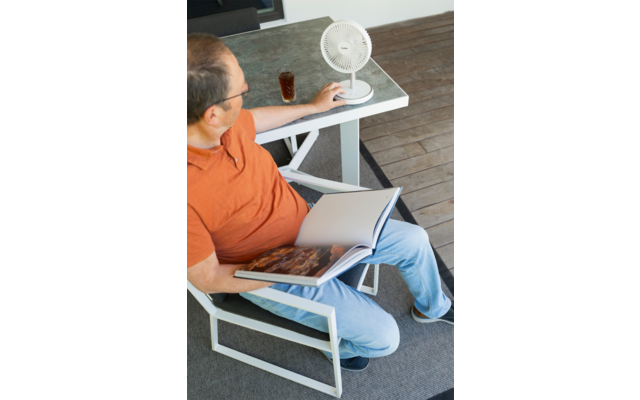 Personne assise sur une chaise extérieure avec haut orange et jean, tenant un livre ouvert sur les genoux ; sur la table à côté un petit ventilateur blanc et un verre contenant une boisson foncée