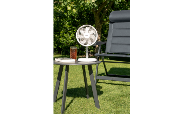Petit ventilateur blanc posé sur une table d'appoint ronde dans un jardin, à côté d'un verre de boisson glacée et d'un fauteuil d'extérieur gris foncé ; gazon et arbres en arrière-plan