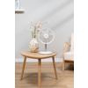 Petit ventilateur de table blanc sur une table d'appoint ronde en bois, à côté d'un vase clair avec des fleurs ; un accoudoir de fauteuil et un rebord de fenêtre visibles en arrière-plan