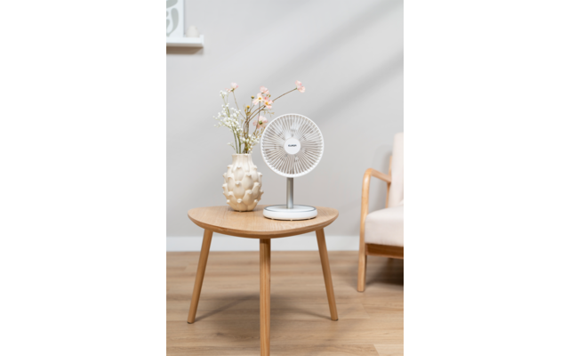 Petit ventilateur de table blanc sur une table d'appoint ronde en bois, à côté d'un vase clair avec des fleurs ; un accoudoir de fauteuil et un rebord de fenêtre visibles en arrière-plan