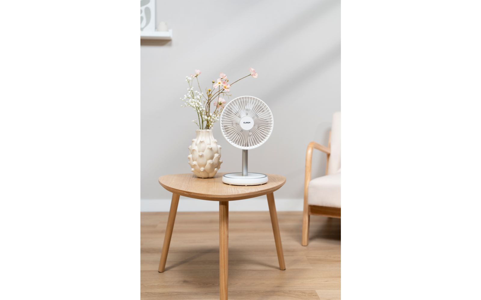 Petit ventilateur de table blanc sur une table d'appoint ronde en bois, à côté d'un vase clair avec des fleurs ; un accoudoir de fauteuil et un rebord de fenêtre visibles en arrière-plan