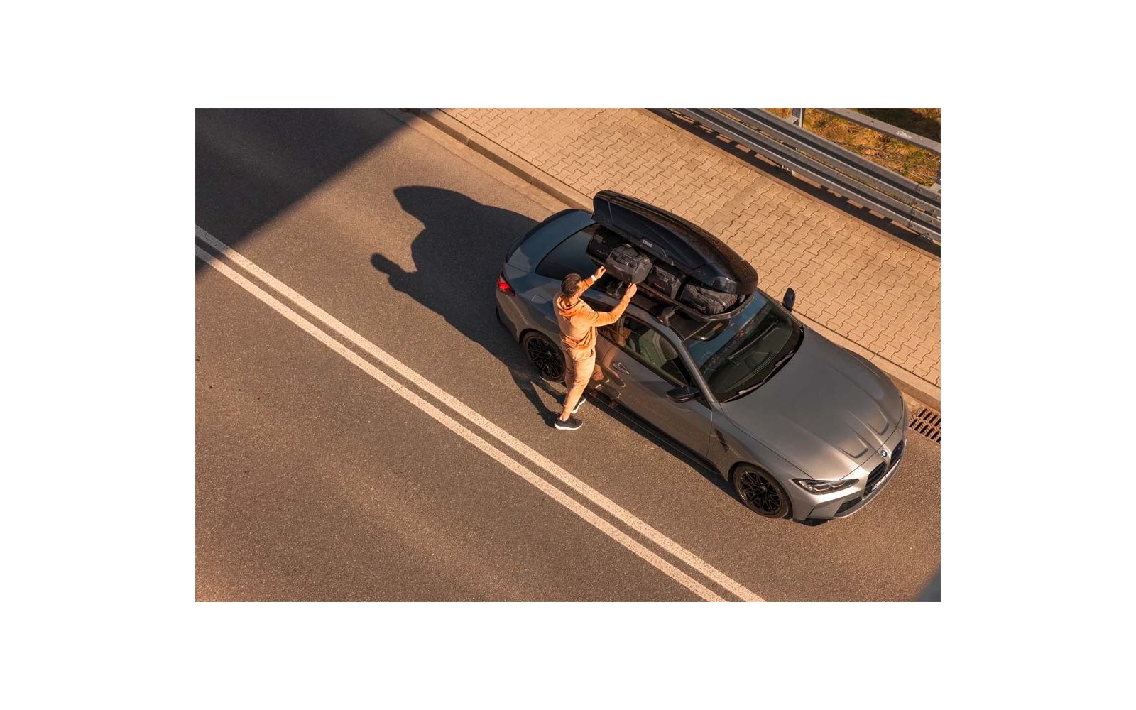 Graues Auto auf einer Straße mit offenem Dachkoffer; Person in orangefarbener Kleidung legt einen Gegenstand hinein; weiße Fahrbahnmarkierungen und Gehweg mit Leitplanke sichtbar