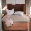 Bett mit beigefarbenem Spannbettlaken, zwei weißen Kissen, hellbeige Jacke an der Ecke, Holzbettgestell, Hängeschrank und Nachttische mit Lampe