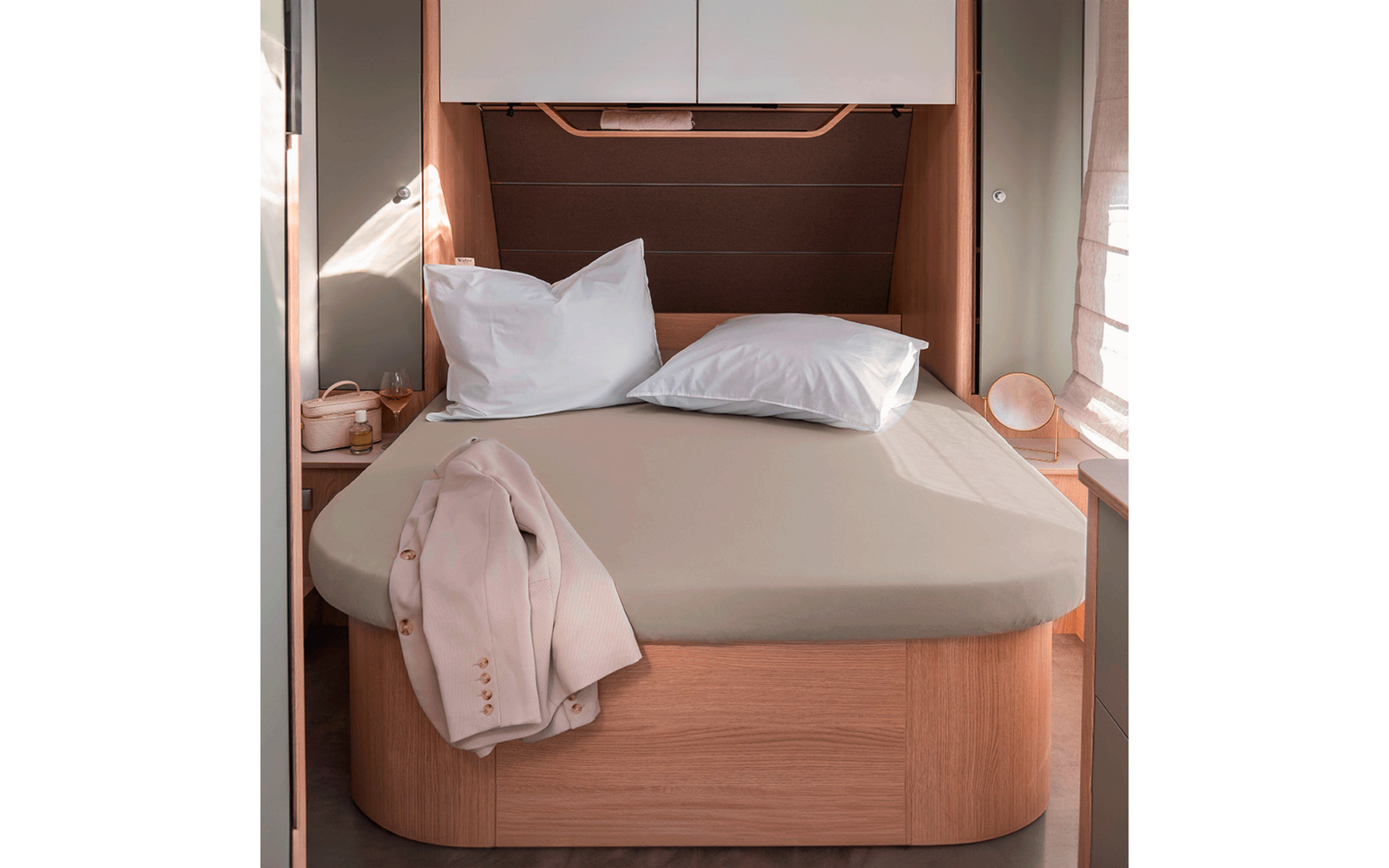 Bett mit beigefarbenem Spannbettlaken, zwei weißen Kissen, hellbeige Jacke an der Ecke, Holzbettgestell, Hängeschrank und Nachttische mit Lampe